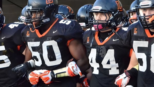 Un grupo de jugadores de fútbol de The Linsly School está en uniformes negros y naranjas con cascos en el campo.
