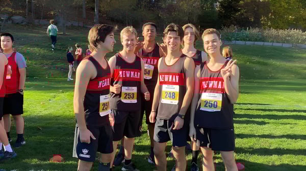 Un grupo de jóvenes atletas está en el campo de deportes de The Newman School con árboles y otros corredores al fondo.