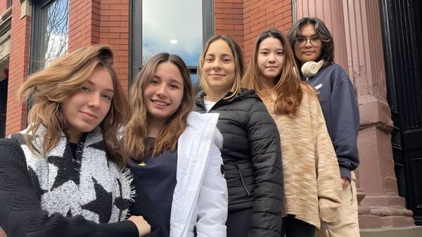 Un grupo de jóvenes mujeres está de pie frente a un edificio de ladrillo en el campus de The Newman School y están conversando.