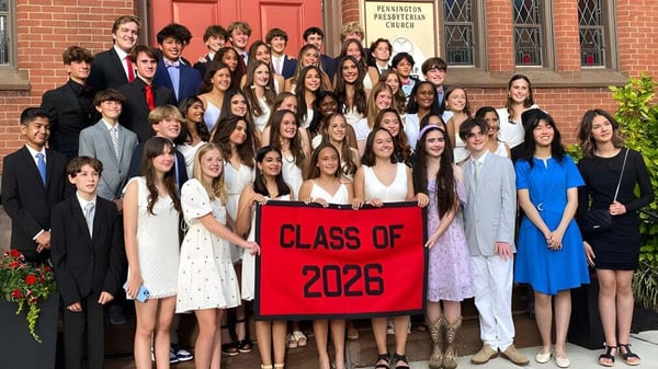Un grupo de estudiantes de The Pennington School está vestido de manera formal frente a un edificio de ladrillo con una pancarta de la clase 2026.