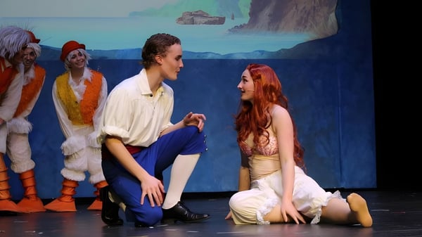 Una representación teatral muestra a una pareja en el escenario frente a un telón de fondo con paisaje oceánico en el campus de The Vanguard School.