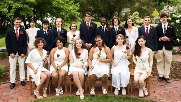 Un grupo de estudiantes de la The White Mountain School posan juntos vestidos formalmente en un área de jardín con vegetación verde y muros de ladrillo.
