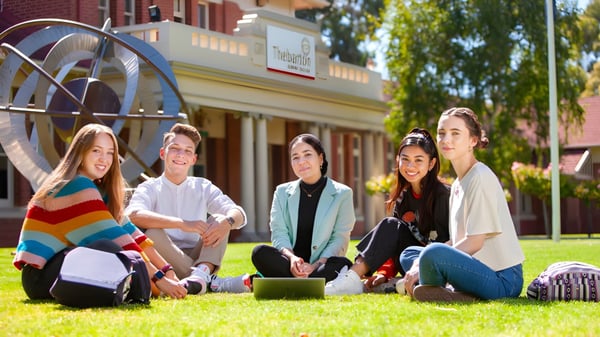 Un grupo de estudiantes está sentado en la hierba frente a un edificio de ladrillo en el campus del Thebarton Senior College.