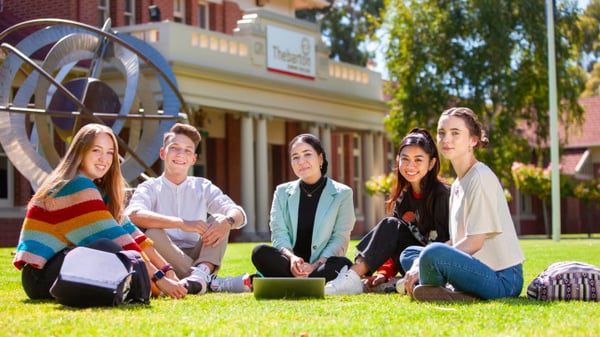 Un grupo de estudiantes está sentado en el prado frente a un edificio de ladrillo con una gran rueda en el terreno del Thebarton Senior College.