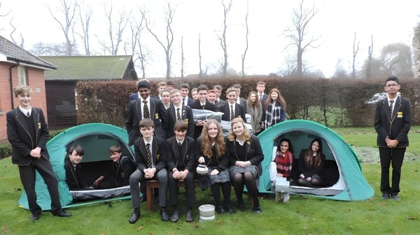 Un grupo de estudiantes de la Thetford Grammar School posan al aire libre frente a una cabaña de madera y árboles.