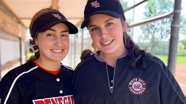 Dos alumnas sonrientes de la Thomas Haney Secondary School con gorras de béisbol y uniforme están de pie frente a un edificio.
