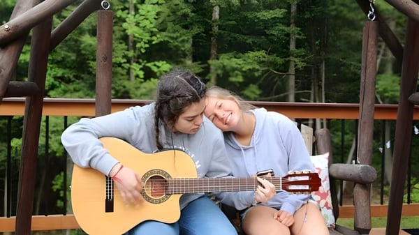 Dos estudiantes de Thornton Academy están sentadas en un banco de madera en el bosque, una toca la guitarra, la otra escucha.
