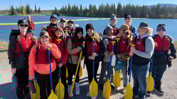Un grupo de estudiantes de la Three Hills School está con chalecos salvavidas rojos y amarillos frente a un lago rodeado de pinos.