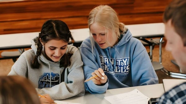 Dos estudiantes de la Three Rivers Christian School trabajan juntas en un escritorio con un fondo de revestimiento de madera.