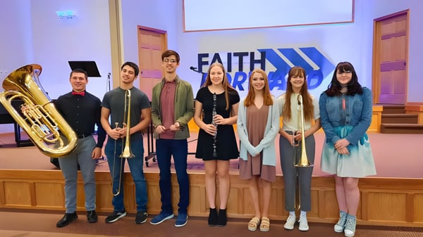 Un grupo de estudiantes de la Thunder Ridge High School está con instrumentos musicales frente a un gran cartel de Faith en un espacio de iglesia.