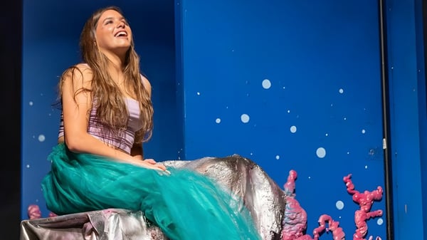 Una joven mujer está sentada en un escenario frente a un fondo azul con puntos blancos en el campus de la Tilton School.