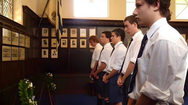 Estudiantes de la Timaru Boys School están frente a una pared con fotografías enmarcadas y un arreglo floral.