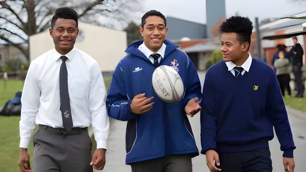 Tres estudiantes de la Timaru Boys School en uniforme escolar con un balón de rugby frente a un edificio escolar y árboles.
