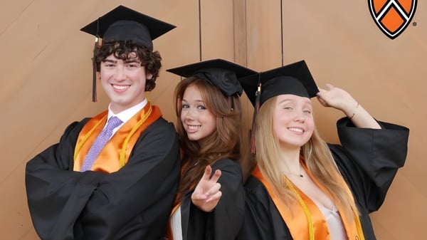Tres graduados de TMI Episcopal posan con birretes y togas de graduación frente a una pared de madera con el logo de la escuela.