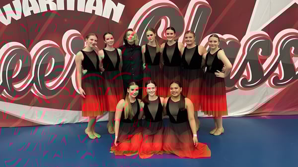 Un grupo de jóvenes estudiantes del Tommy Douglas Collegiate está en trajes coordinados en negro y rojo frente a un gráfico rojo y blanco como grupo de baile.