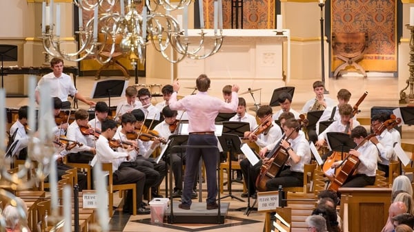 La orquesta de la Tonbridge School toca en un escenario con magníficos candelabros y arquitectura decorativa de fondo.