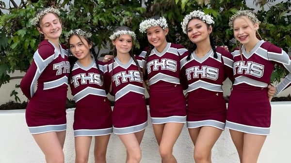 Un grupo de estudiantes de la Torrance High School en uniformes de animación con coronas de flores posan al aire libre frente a un fondo verde.