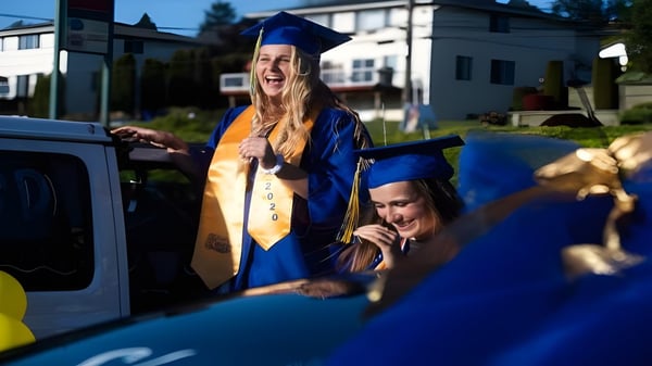 Una graduada sonriente en toga azul está al lado de un auto en el campus del Torrance Unified School District con otros graduados.