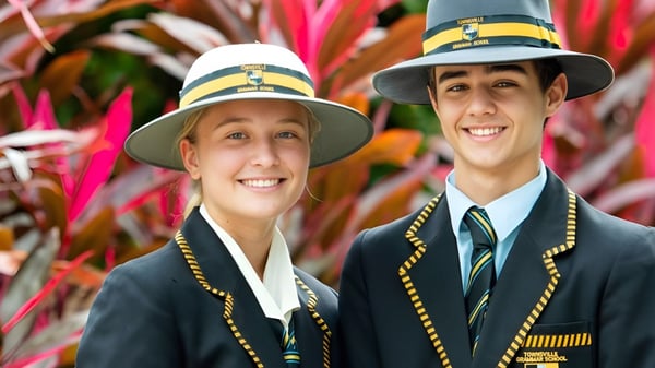 Dos estudiantes de la Townsville Grammar School llevan sombreros tradicionales y uniformes frente a un fondo colorido.