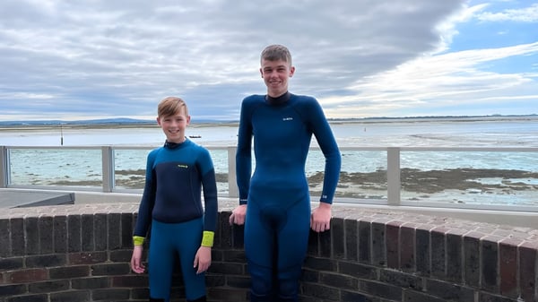 Dos personas en trajes de neopreno azules están de pie junto a una pared con vista a un cuerpo de agua frente a montañas en el campus de la Trafalgar School.