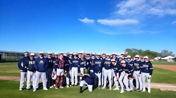 Un grupo de jugadores de béisbol está en el campo de la Tri-Cities Prep Catholic High School bajo un cielo despejado.