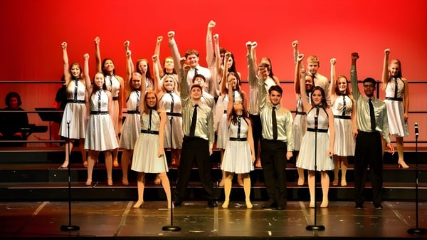 Estudiantes de la Tri-State Christian School están de pie en trajes blancos en el escenario y levantan los brazos frente a un fondo rojo.