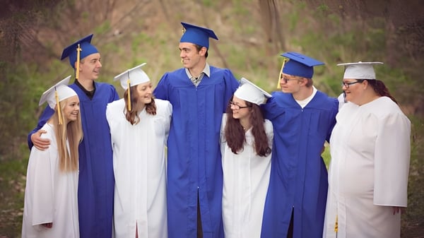 Un grupo de graduados en togas azules y blancas está de pie en el área exterior de la Tri-State Christian School frente a un fondo boscoso.
