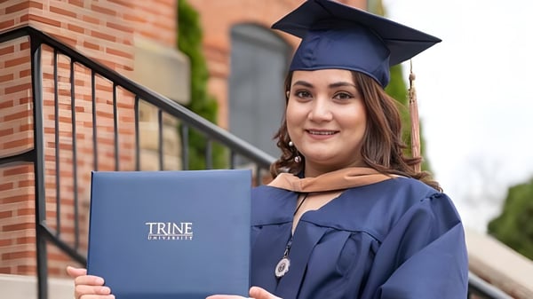 Una graduada sonriente de la Trine University se encuentra frente a un edificio de ladrillo y sostiene su diploma en la mano.