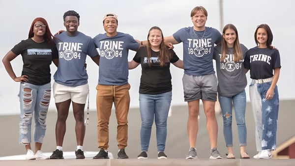 Un grupo de estudiantes se encuentra frente a un cielo nublado con camisetas de la Trine University.