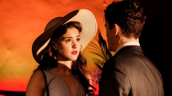 Una mujer con sombrero de ala ancha y un hombre en traje están en un diálogo intenso frente a un fondo naranja en el campus de la Tring Park School for the Performing Arts.