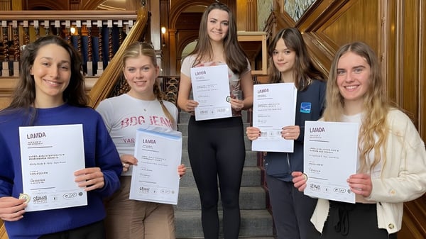 Cinco estudiantes de la Tring Park School for the Performing Arts sostienen sus diplomas frente a la cámara en un espacio histórico.