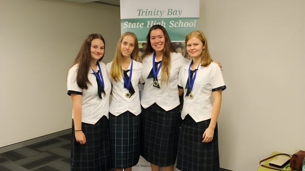 Cuatro estudiantes en uniformes escolares están juntas frente al cartel de la Trinity Bay State High School.