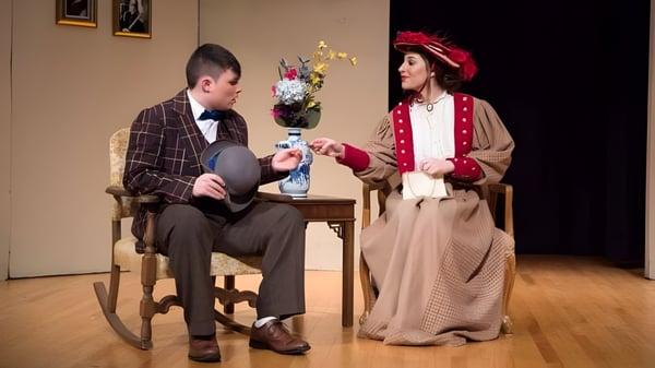 Un hombre en una mecedora y una mujer con un jarrón de flores están vestidos con trajes históricos en el campus de la Trinity Catholic Jr/Sr High School.