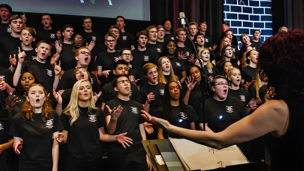 Un gran grupo de estudiantes de la Trinity Catholic High School está en un escenario frente a una pared de ladrillo.