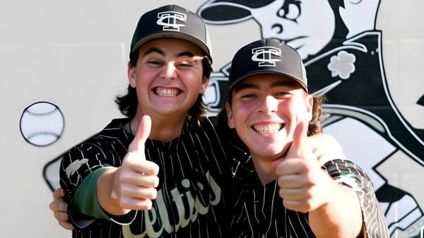 Dos estudiantes de la Trinity Catholic High School llevan gorras de béisbol y camisetas y muestran el pulgar hacia arriba frente a un mural con un personaje de cómic.