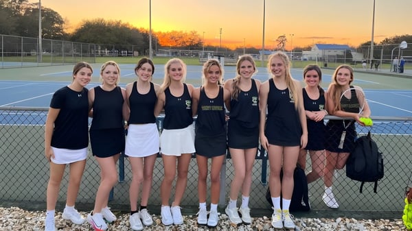 Un grupo de jóvenes estudiantes de tenis posan en la cancha de tenis de la Trinity Catholic High School al atardecer.
