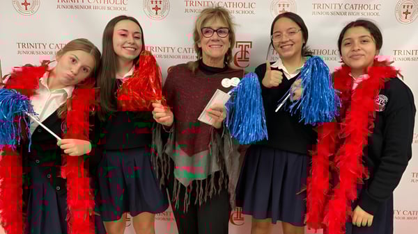 Un grupo de estudiantes con accesorios de plumas de colores está frente al logo de la Trinity Catholic Jr/Sr High School.