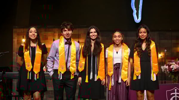 Un grupo de cinco alumnas y alumnos está en ropa de graduación con velas y flores de fondo en el campus de la Trinity Christian Academy.