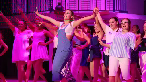 Un grupo de alumnas y alumnos baila en el escenario del Trinity College frente a una iluminación vibrante en tonos rosa y violeta.