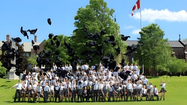 Un grupo de graduados y graduadas en togas y birretes blancos se reúne en el césped de la Trinity College School.