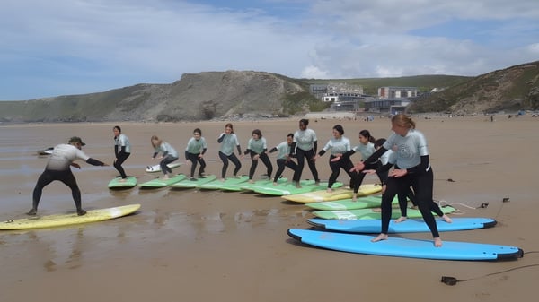 Las alumnas de la Truro High School for Girls están de pie con trajes de neopreno sobre tablas de surf en la playa frente a montañas y edificios.