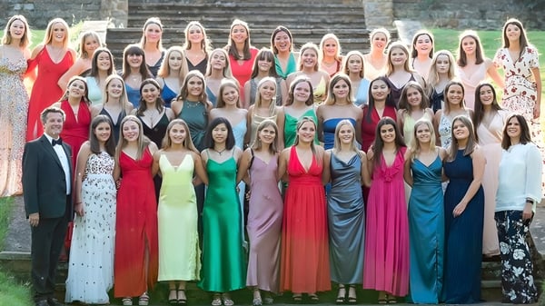 Un gran grupo de mujeres con vestidos coloridos está de pie frente a un edificio de piedra en el campus de Tudor Hall School.