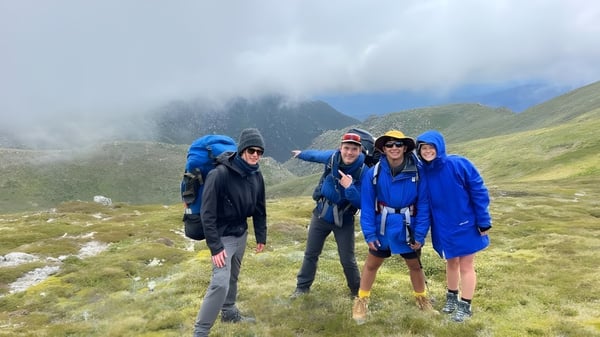 Un grupo de estudiantes de la UC-Lake Ginninderra está en chaquetas de lluvia coloridas en un sendero montañoso neblinoso con altas cumbres al fondo.