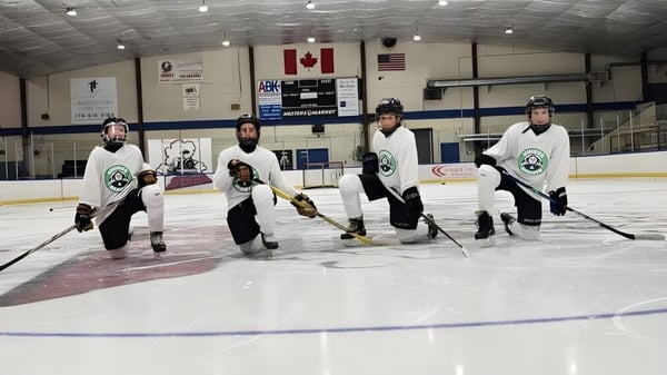 Un grupo de jugadores de hockey en uniforme está en la pista de hielo de la Unisus School con la bandera canadiense y americana de fondo.