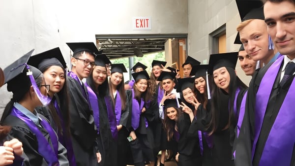 Graduadas y graduados de la University Hill Secondary School están juntos en un pasillo con togas violetas.