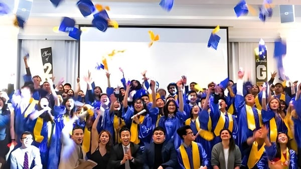Estudiantes de la Urban International High School celebran su ceremonia de graduación en togas azules y amarillas con los brazos en alto.