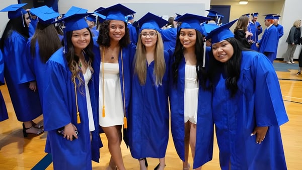 Un grupo de jóvenes en túnicas de graduación azules están juntas en el campus del Urbandale Community School District.