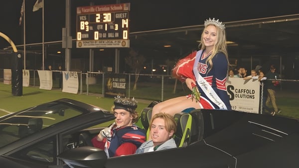 Una mujer con corona y banda está de pie junto a un auto frente a un tablero de anuncios en el campus de la Vacaville Christian School.