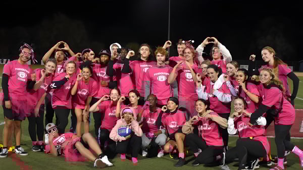 Un gran grupo de jóvenes mujeres en camisetas rosas uniformes está de pie afuera por la noche en el campus de la Vacaville Christian School.