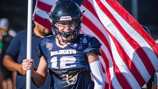 Un jugador de football de la Valencia High School sostiene una bandera americana frente a un fondo de estadio.